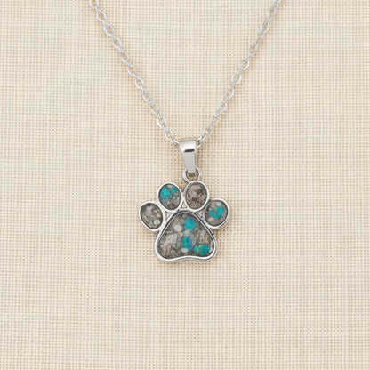 Silver paw print necklace with gemstones on a white surface with pink flowers in the background